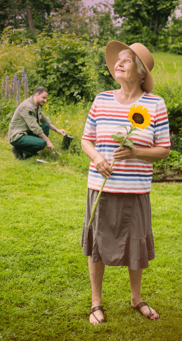 Eine alte Frau steht in der Mitte ihres Gartens mit einer Sonnenblume in der Hand und blickt in den Himmel, während im Hintergrund der Gärtner das Blumenbeet in Ordnung bringt.