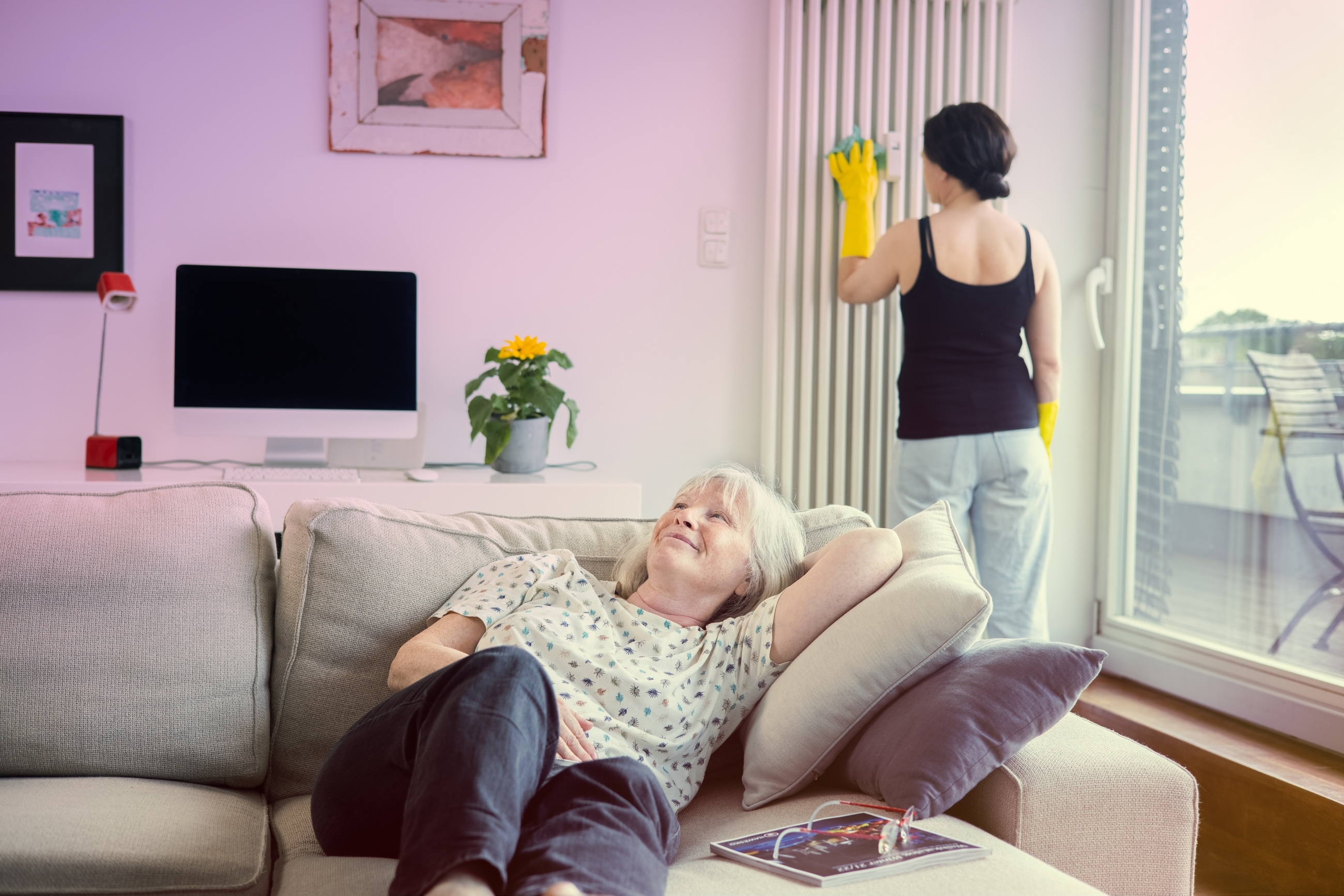 Eine ältere Frau entspannt sich auf dem Sofa, während im Hintergrund des Bildes eine Putzfrau einen Heizkörper reinigt.